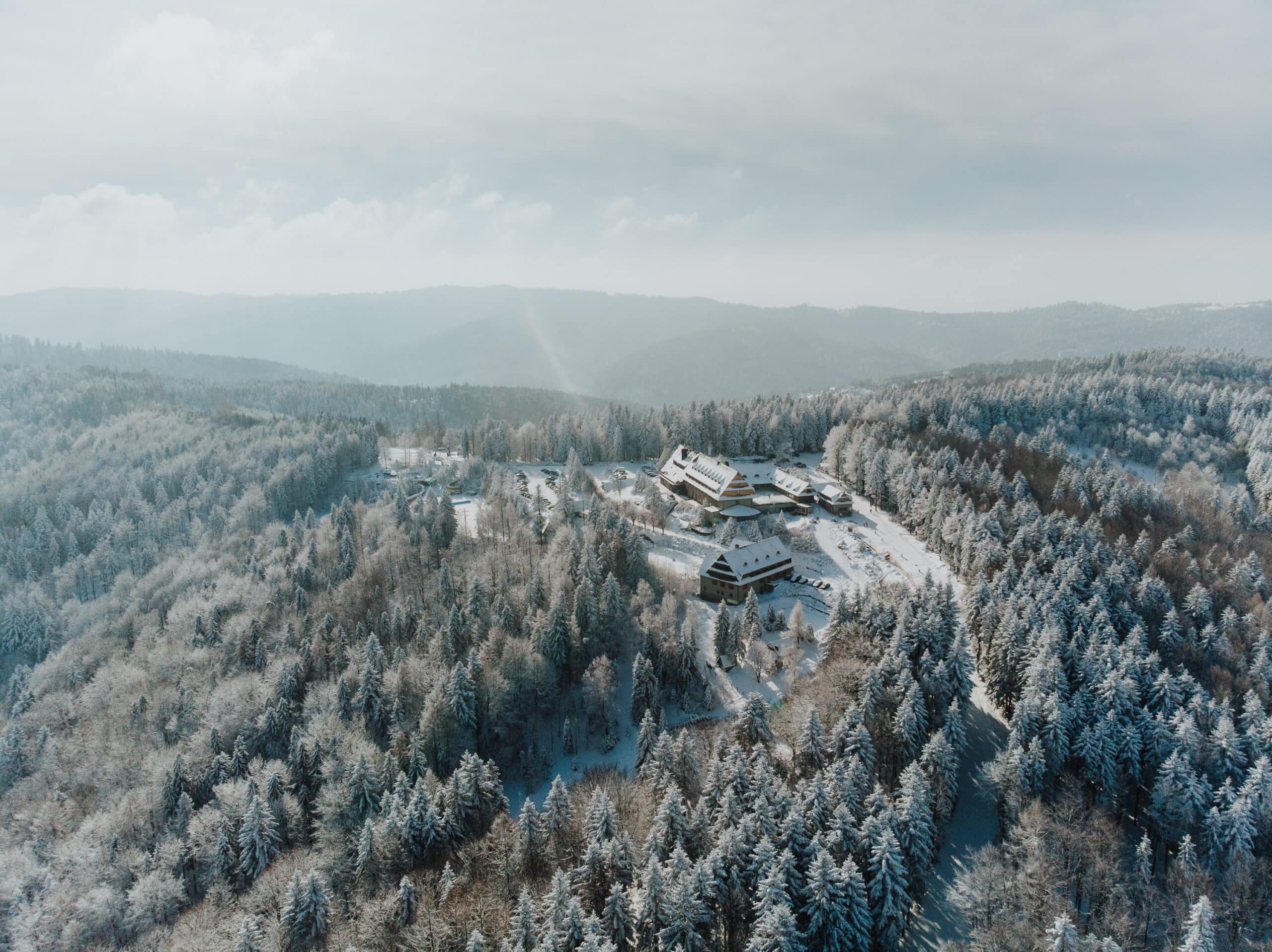 Aerial view of a snow-covered forest and chalet-style buildings in a mountainous landscape.